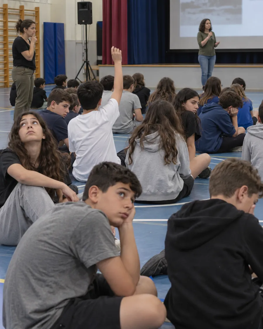 טקס באולם ספורט: תלמידים יושבים על הרצפה ומשתעממים, מורה מבקשת שקט
